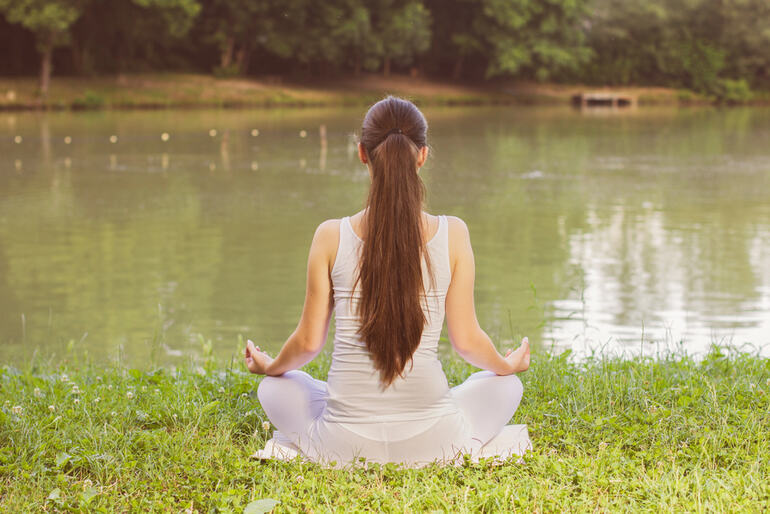 A woman is meditating by a lake, considering the benefits of meditation