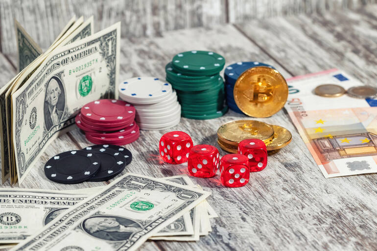 Casino equipment, including poker chips, U.S. dollar bills, red dice, and cryptocurrency coins on a wooden table