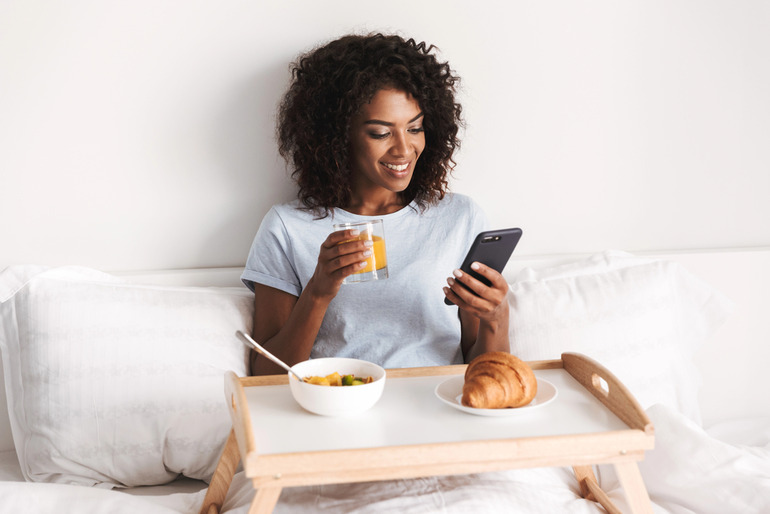 Woman sitting in bed with breakfast and juice, smiling while looking at her phone, highlighting morning screen time habits.