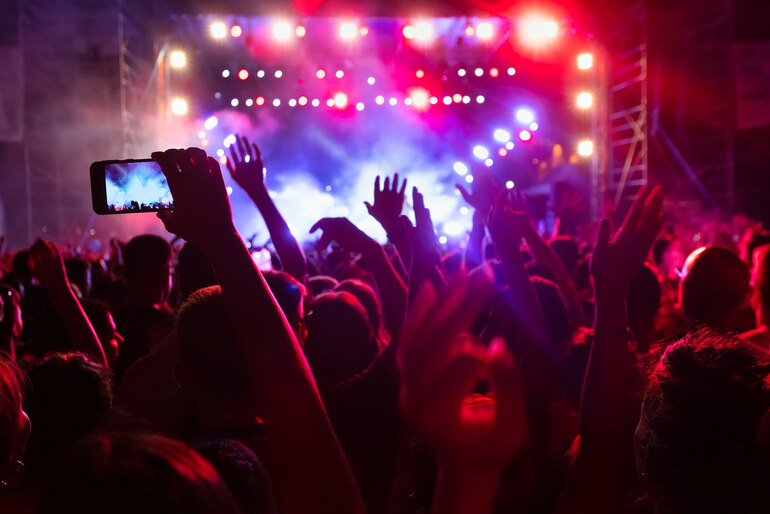 Crowd at a music festival with raised hands and a person recording the stage on a smartphone under vibrant concert lighting