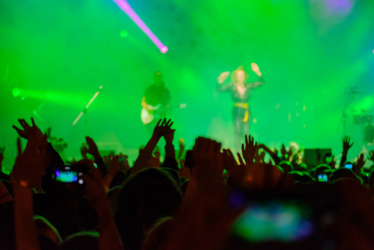 Crowd dancing and raising hands at a live concert with a guitarist performing on stage under vibrant purple lights