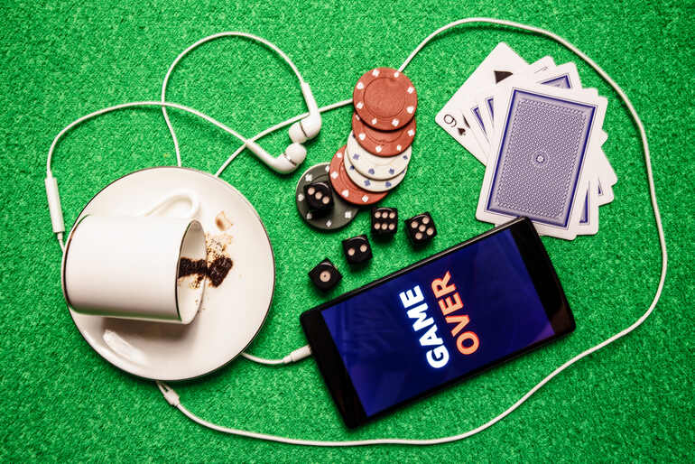 A smartphone displaying "GAME OVER" lies on a green felt surface surrounded by poker chips, dice, and playing cards
