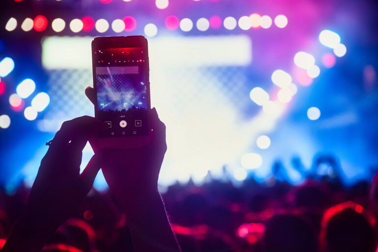 A person records a live concert performance with their smartphone, capturing the vibrant stage lights and a cheering crowd in the background.