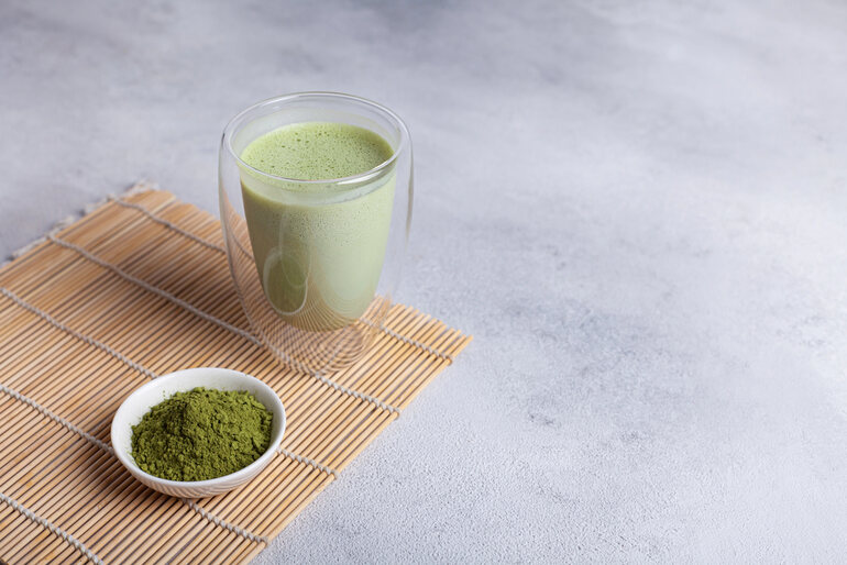 A glass of matcha and a bowl of kratom powder on a wooden mat.