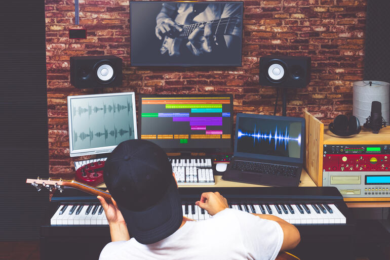 Musician in a home recording studio working on music production with a guitar, keyboard, and multiple screens