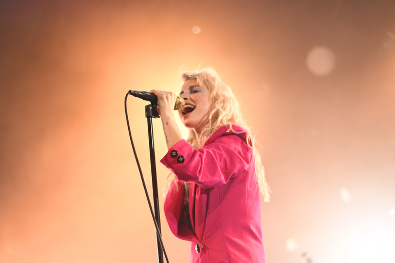 Female singer passionately performing live on stage in a vibrant pink outfit with dramatic lighting