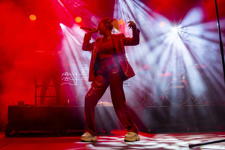 A singer performing energetically on stage under red and white lights during a live concert.