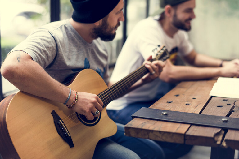 Two men sitting at a wooden table, one playing an acoustic guitar while the other listens and smiles during a casual music session.