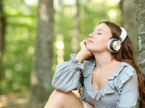 Woman sitting outdoors in a forest with headphones on, eyes closed, enjoying calming music and relaxation.