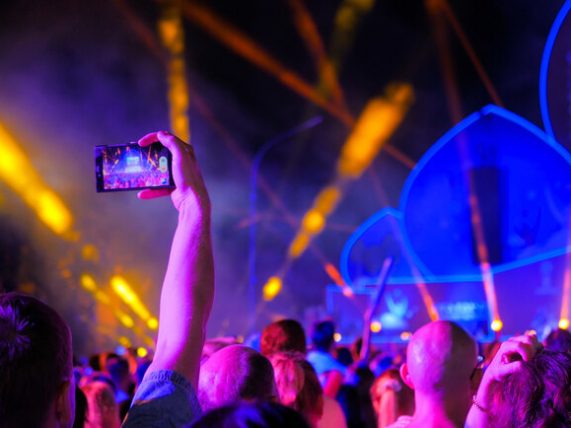 Crowd at a night music festival with neon lights and lasers as a fan films the digital-inspired stage on a smartphone, echoing gaming culture’s impact on live music.