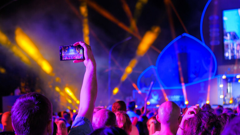 Crowd at a night music festival with neon lights and lasers as a fan films the digital-inspired stage on a smartphone, echoing gaming culture’s impact on live music.