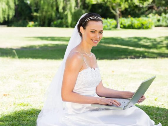 Smiling bride in a wedding dress using a laptop outdoors, symbolizing online matchmaking and international marriage topics.