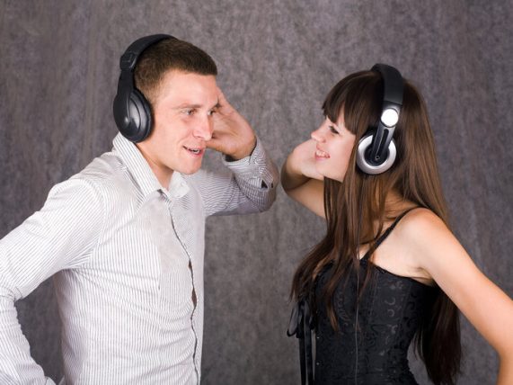 A man and woman wearing headphones facing each other, smiling as they share music together, suggesting chemistry and connection.