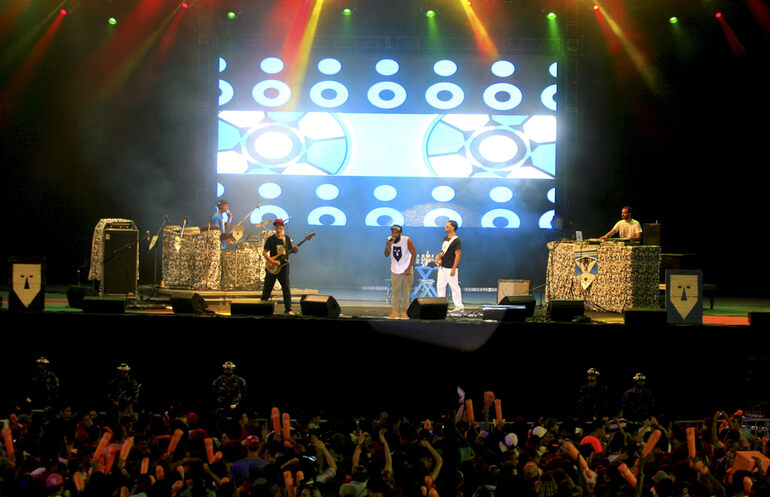 Hip-hop group performing on stage with DJs, musicians, and a large crowd at a live concert.