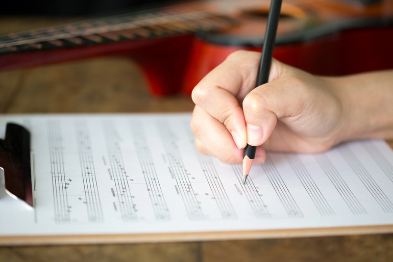 Musician writing song lyrics or composing music on sheet paper with a guitar in the background, representing ghostwriting in the music industry.