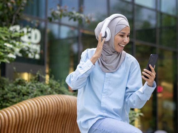 The picture of a smiling Pakistani woman with headphones sitting in the foreground engages in listening to music on her smartphone- this depicts the ease of seeing the Melbet app.