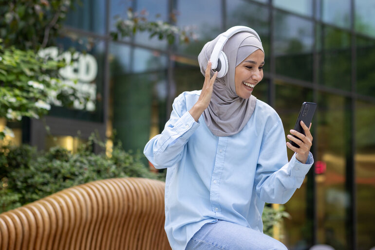 The picture of a smiling Pakistani woman with headphones sitting in the foreground engages in listening to music on her smartphone- this depicts the ease of seeing the Melbet app.