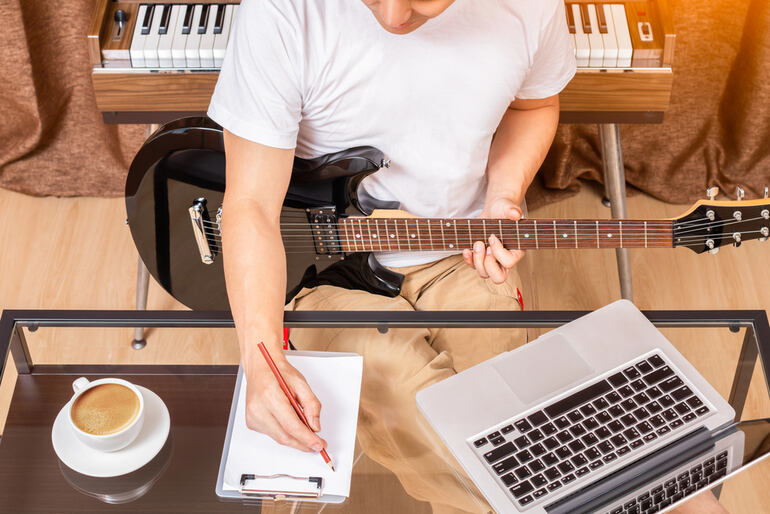 Aspiring musician composing music at home with an electric guitar, laptop, notebook, and keyboard, representing modern and compact music creation solutions.