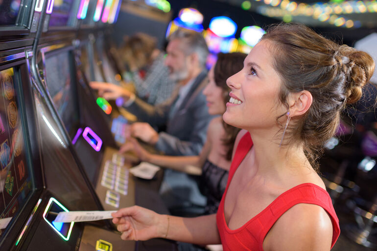 A smiling woman in a red dress inserts a ticket into a glowing slot machine in a vibrant casino