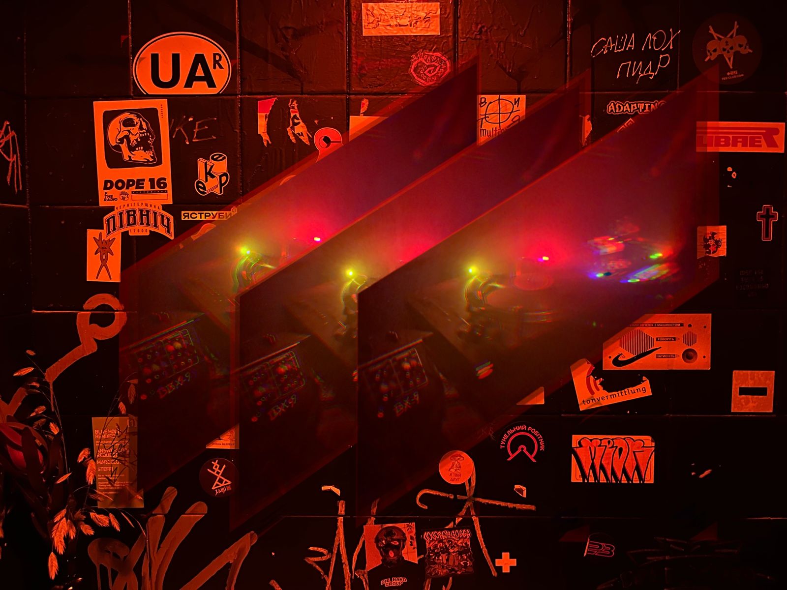 A dark, atmospheric scene bathed in red light, featuring a wall covered in stickers and graffiti behind three translucent, overlapping glass panels.