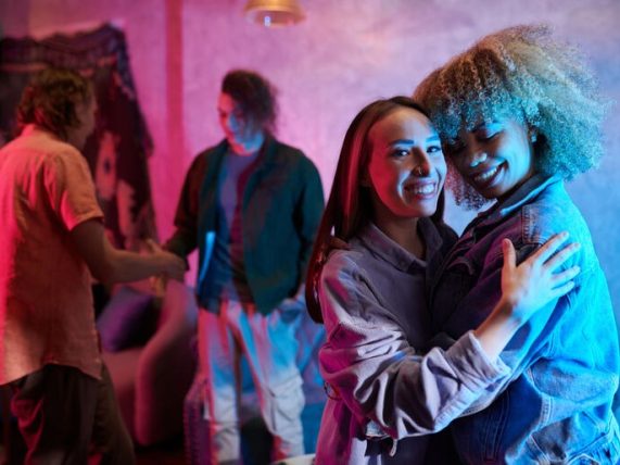 Two young women embracing and smiling at a party with moody, purple and blue Euphoria-style neon lighting, with other guests blurred in the background.