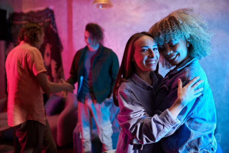 Two young women embracing and smiling at a party with moody, purple and blue Euphoria-style neon lighting, with other guests blurred in the background.