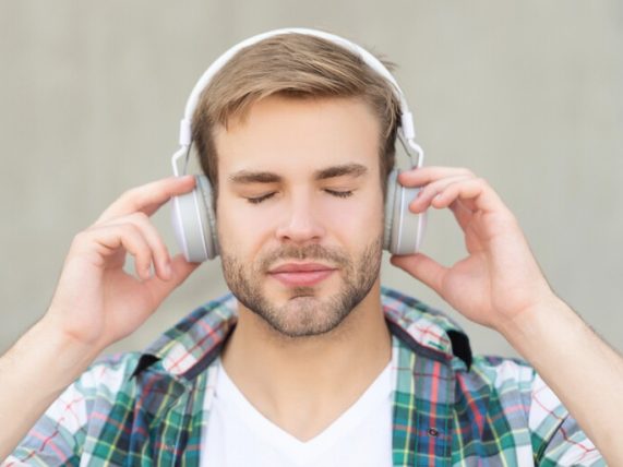 A close-up of a young man with his eyes closed, deeply focused and listening intently to socially critical music through large white headphones. 