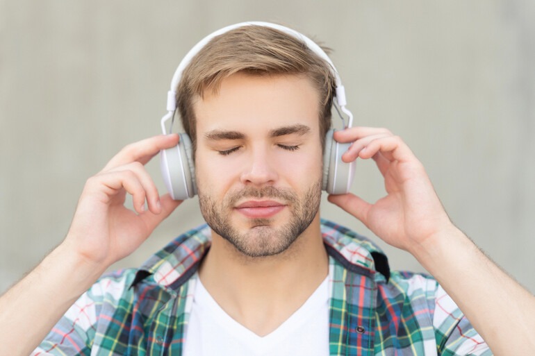 A close-up of a young man with his eyes closed, deeply focused and listening intently to socially critical music through large white headphones.