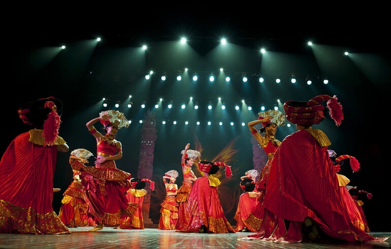 Dancers in elaborate red and gold costumes perform a traditional stage production under bright theater lights, showcasing a memorable cultural experience.