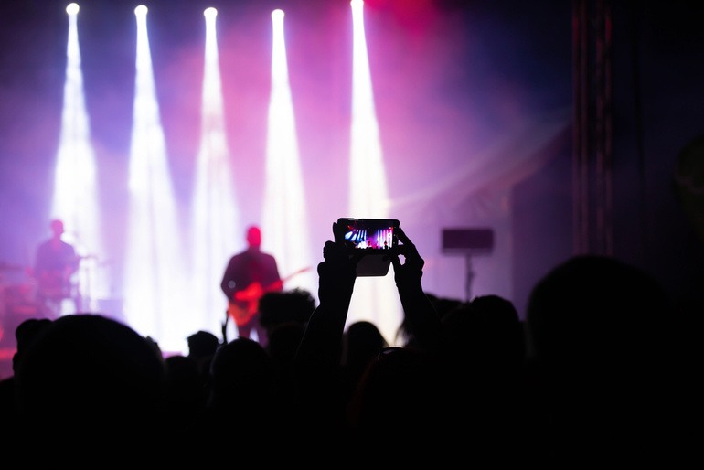 Person filming a live concert on a smartphone in a dark venue with bright stage lights.