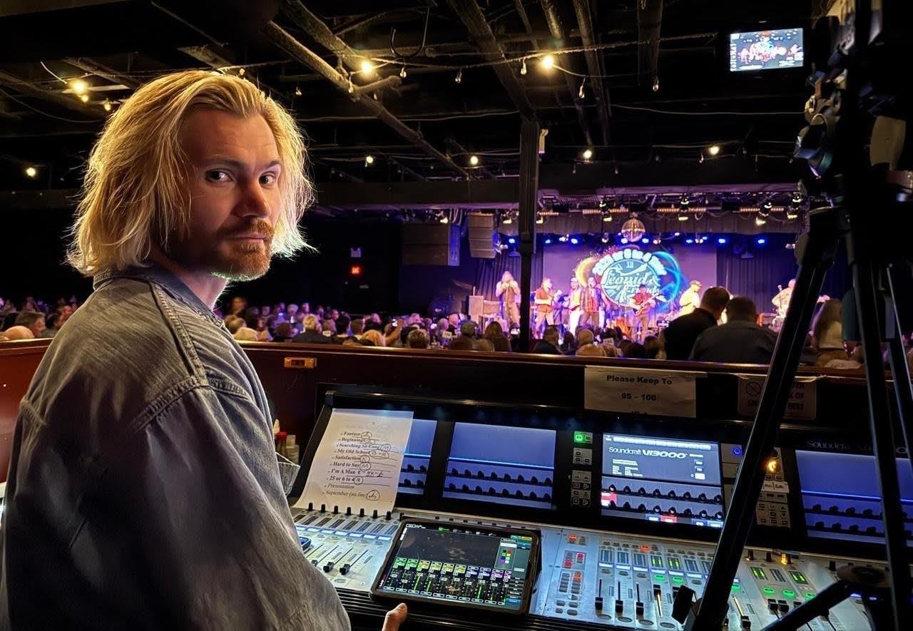 Sound engineer Aleksei Bondarenko at a concert mixing console during a live music performance, with stage lights and musicians in the background.